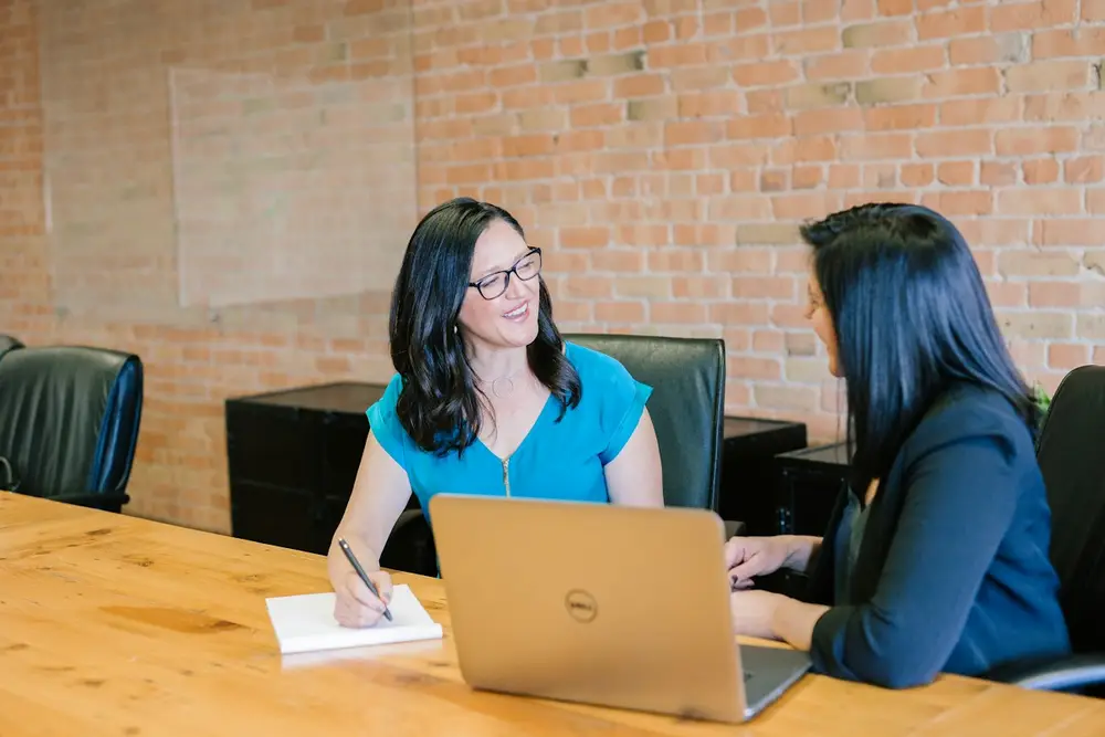 Two colleagues comparing notes at a table with a laptop